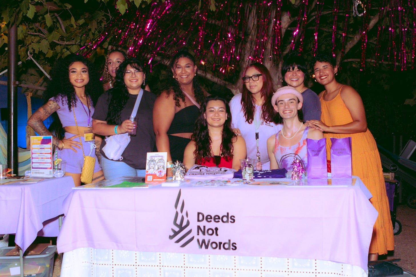 Deeds Not Words team around their organization table at The Canvas of Change outdoor event in Houston, Texas. In the photo, there are nine people standing behind a table with a purple tablecloth with the Deeds Not Words logo. Deeds Not Words team around their organization table at The Canvas of Change outdoor event in Houston, Texas. In the photo, there are nine people standing behind a table with a purple tablecloth with the Deeds Not Words logo.