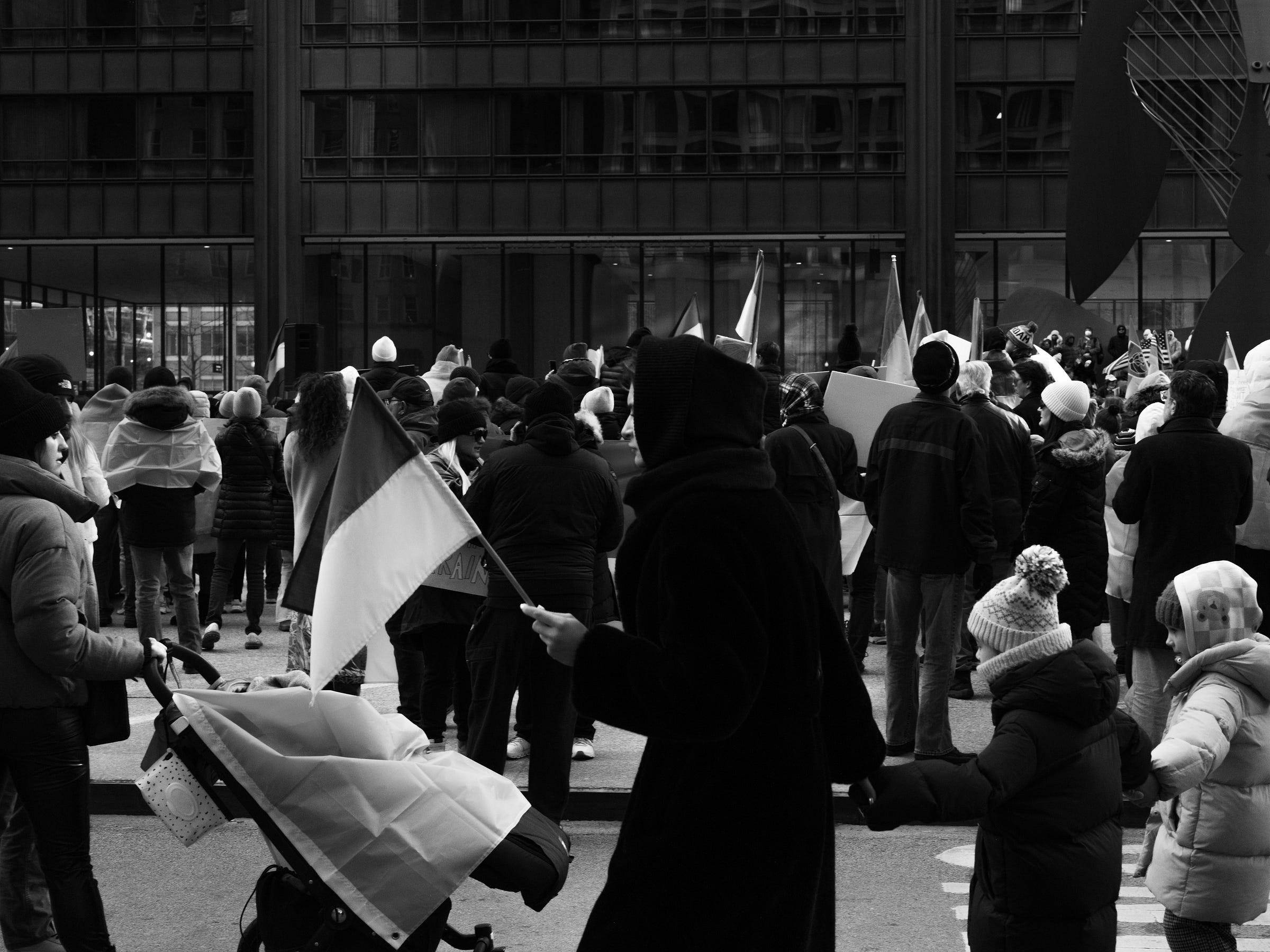 A black and white photograph captures a protest scene with a diverse crowd gathered in front of a modern glass building. A person in a hooded coat waves a small Ukrainian flag while holding hands with a child bundled in winter clothing. A stroller draped with a Ukrainian flag sits nearby. Protesters, many also carrying flags and signs, stand together in solidarity as they listen to speakers addressing the crowd. A black and white photograph captures a protest scene with a diverse crowd gathered in front of a modern glass building. A person in a hooded coat waves a small Ukrainian flag while holding hands with a child bundled in winter clothing. A stroller draped with a Ukrainian flag sits nearby. Protesters, many also carrying flags and signs, stand together in solidarity as they listen to speakers addressing the crowd.