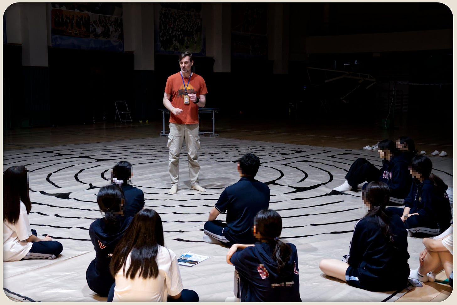 Students sit in a loose circle on a taped labyrinth tarp in a dim school auditorium while the facilitator stands speaking in the center.