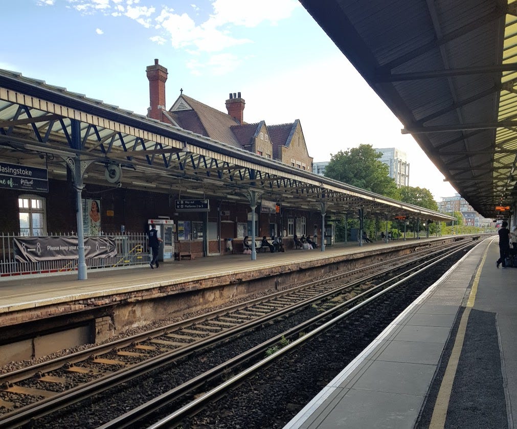 The view from a platform of a UK train station. A few people are on benches on either side of the railway lines. The weather is sunny.