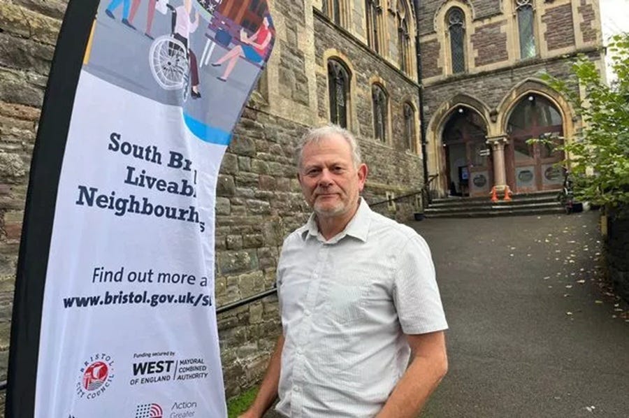Councillor Ed Plowden, a middle-aged white man in a light short-sleeved collared shirt, stands outside a stone church entrance with a Gothic archway. To his left is a vertical banner for the "South Bristol Liveable Neighbourhood" scheme, featuring a partial logo for Bristol City Council and the West of England Combined Authority (WECA).
