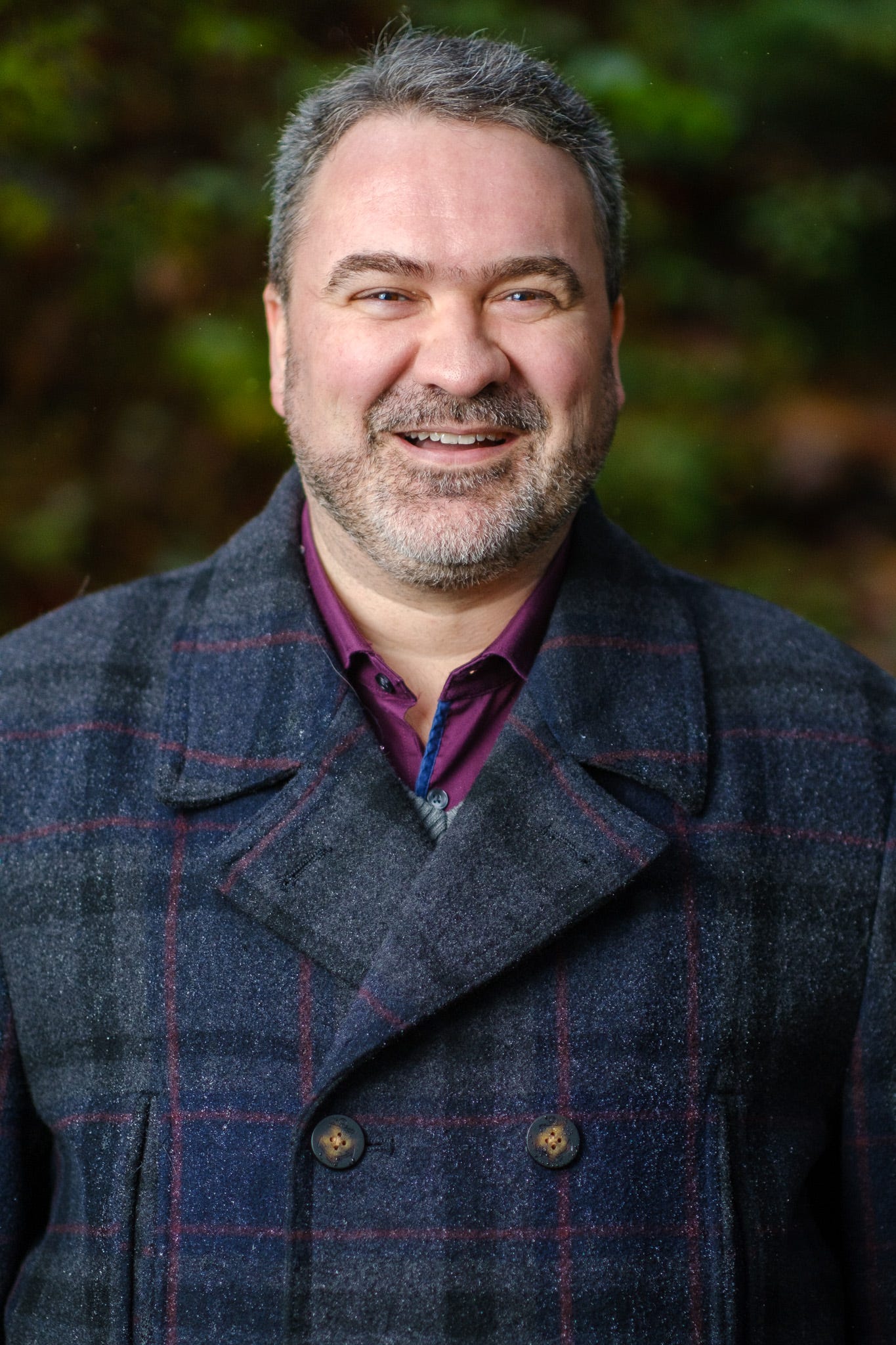Portrait photo of a bearded man smiling, standing against a backdrop of blurry trees.