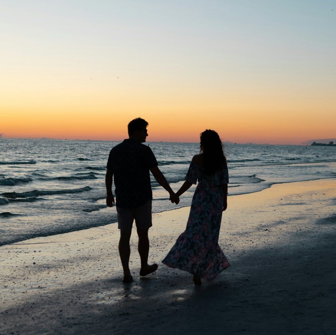 photography of man and woman holding hands each other while walking beside seashore
