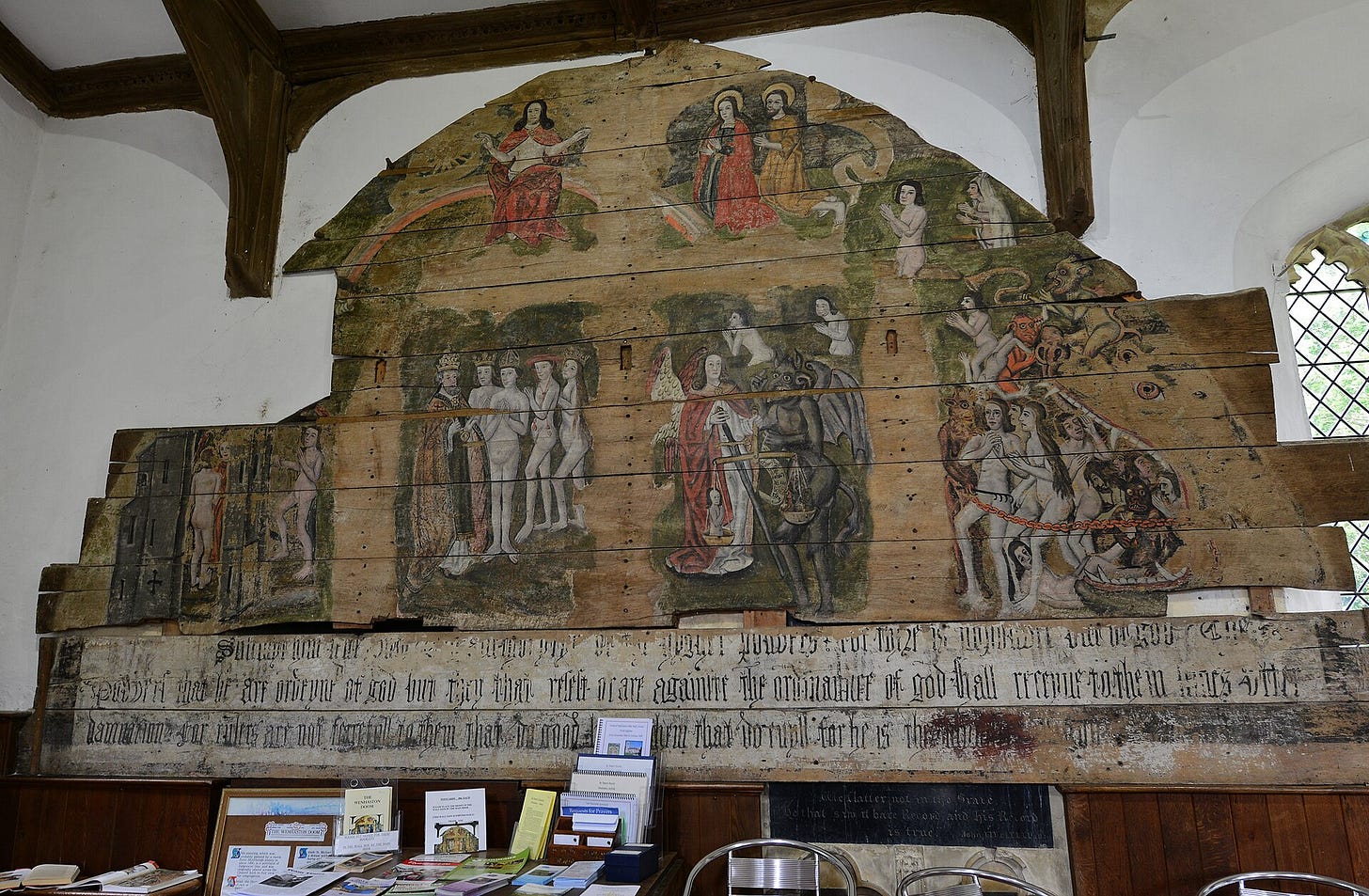 Large painted wooden panel shaped like an arch, hung on a church wall: Christ seated on a rainbow at the top, Saint Michael weighing a soul in the middle, naked figures led toward a gate on the left and into a monster's open mouth on the right, with a line of painted text along the bottom.