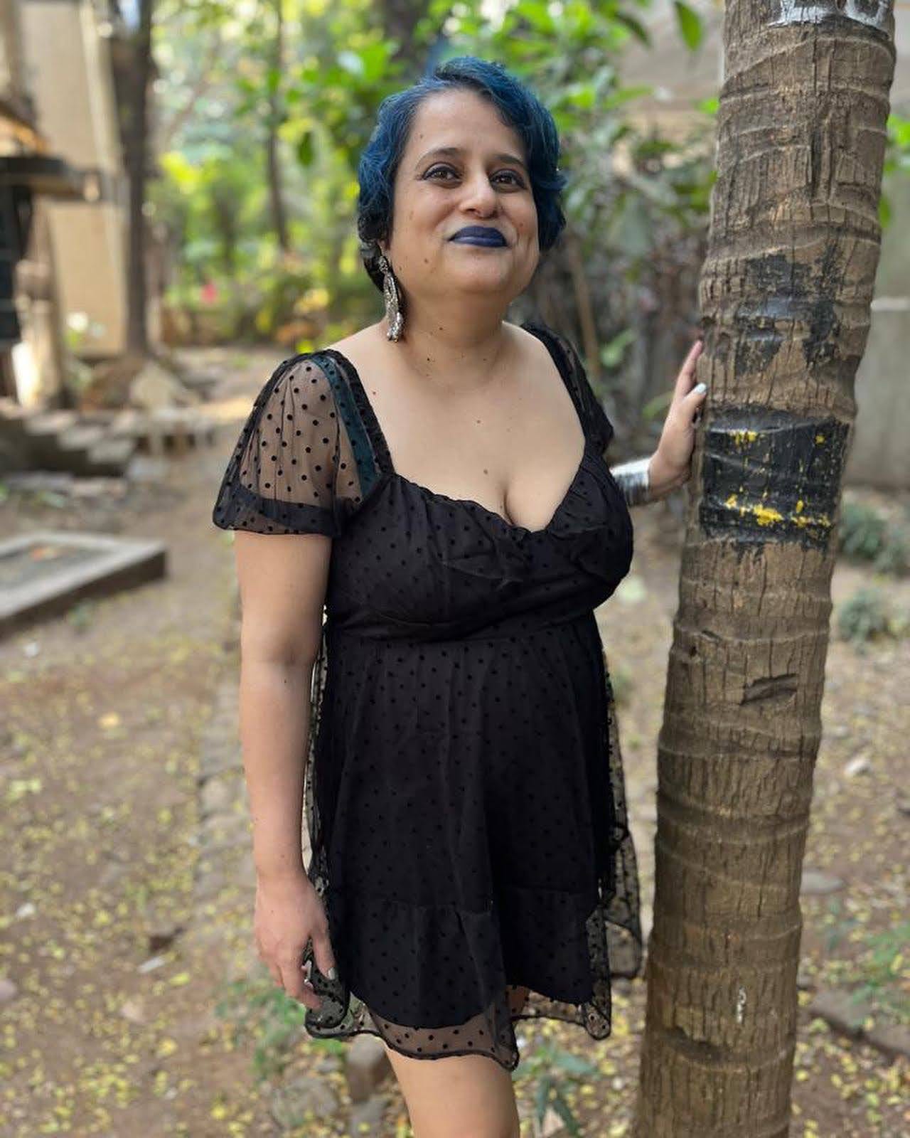 Unmana is standing next to a palm tree. Their hair has blue streaks. They're wearing blue lipstick, grey earrings and a little black dress.