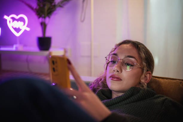 Young woman relaxing on sofa using smartphone in neon-lit room Young woman relaxing on a comfortable sofa, browsing social media on her smartphone in a cozy, neon-lit living room scrolling stock pictures, royalty-free photos & images