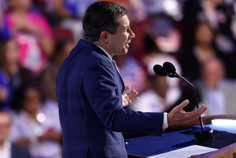 A man in a blue suit jacket, standing at a microphone and gesturing with his hands. A man in a blue suit jacket, standing at a microphone and gesturing with his hands.