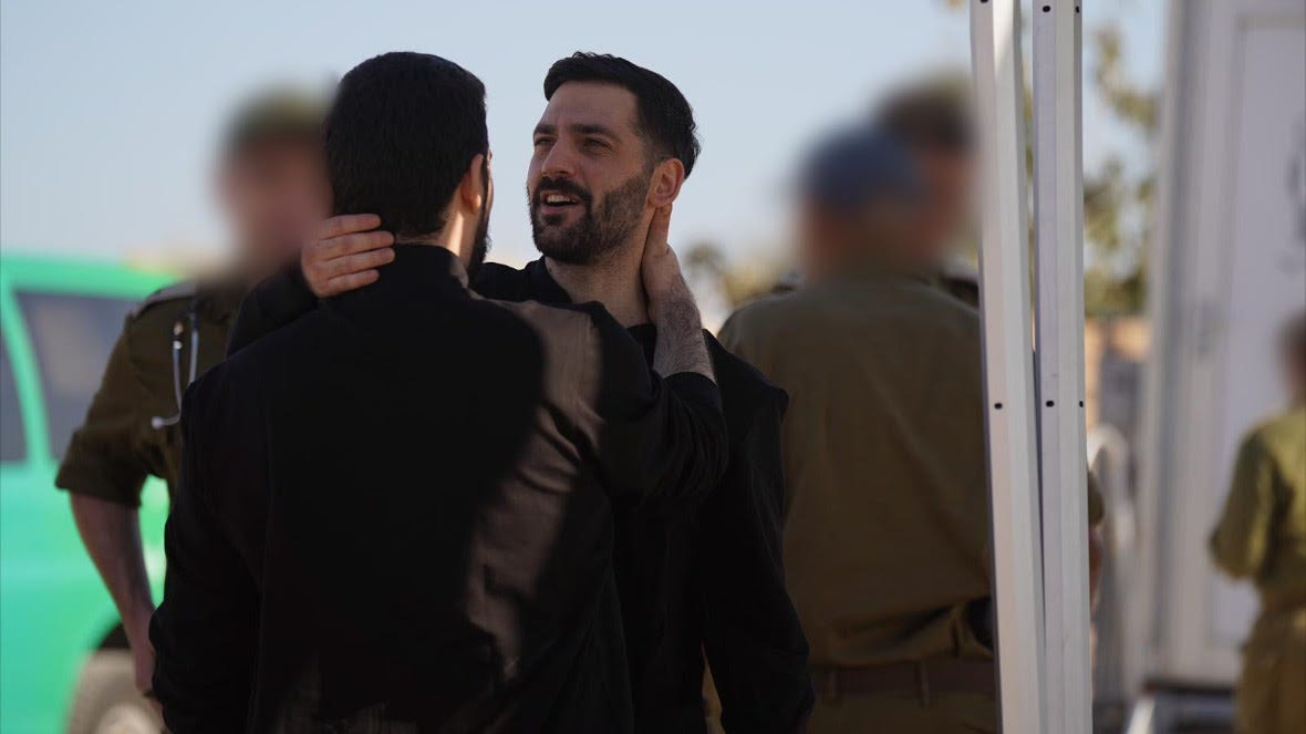 Two men dressed in black are embracing and looking at each other. People dressed in military uniforms are standing in the background, their faces blurred.