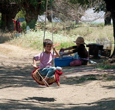 A small, dark-haired child is swinging on a tire. Another boy plays in the background while sitting in the dirt. A small, dark-haired child is swinging on a tire. Another boy plays in the background while sitting in the dirt.