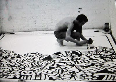 This is a black and white screenshot of a video of an American artist painting himself into a corner in an art gallery, using black and white paint, and he is shirtless as he backs towards the wall. 