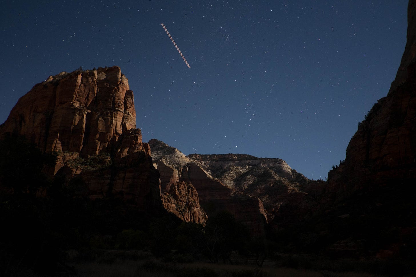A desert canyon at night. Tall red sandstone cliffs rise on the left and right, faintly lit, framing a paler moonlit ridge in the middle distance. A desert canyon at night. Tall red sandstone cliffs rise on the left and right, faintly lit, framing a paler moonlit ridge in the middle distance.