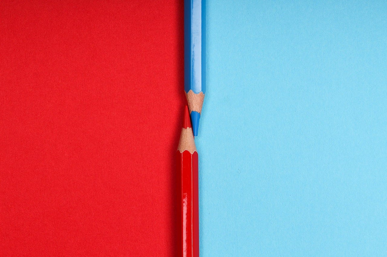 Two sharpened colored pencils - one red and one blue - are placed tip-to-tip on a split background, with red on the left and blue on the right. The red pencil lies on the blue side and the blue pencil on the red side, symbolizing contrast and symmetry."