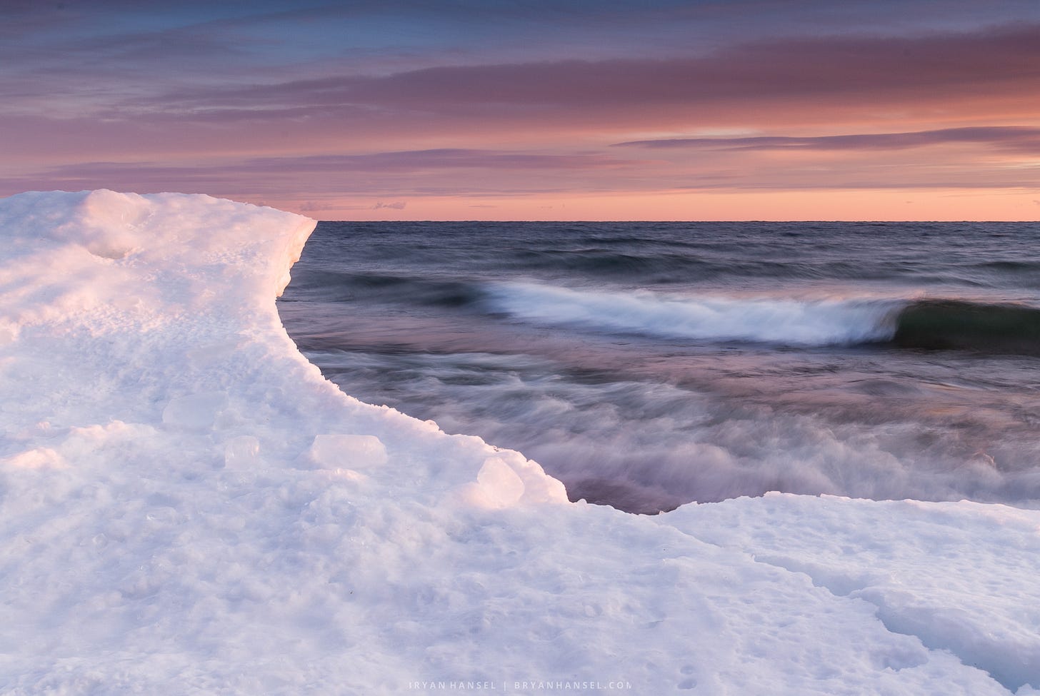An ice formation the shape of a wave is in front of waves rolling in from Lake Superior. At sunset under a pink and orange sky.