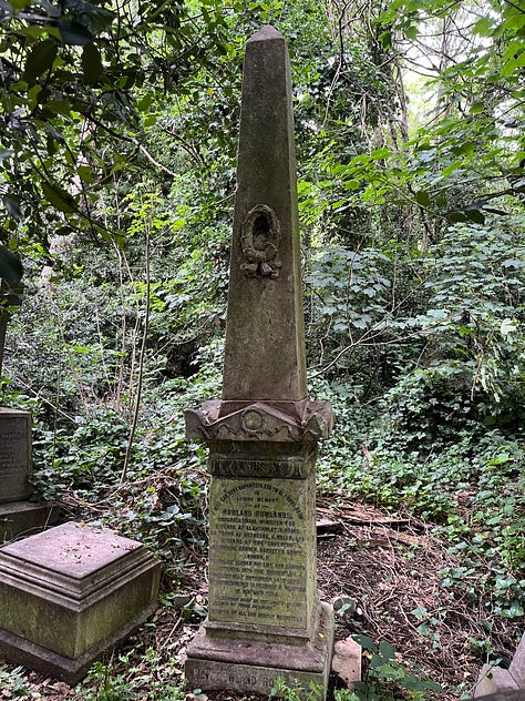 Three obelisk grave monuments with adornments. One has a hand pointing upwards on top, one a laurel wreath carved into the front, and one has an urn on top.