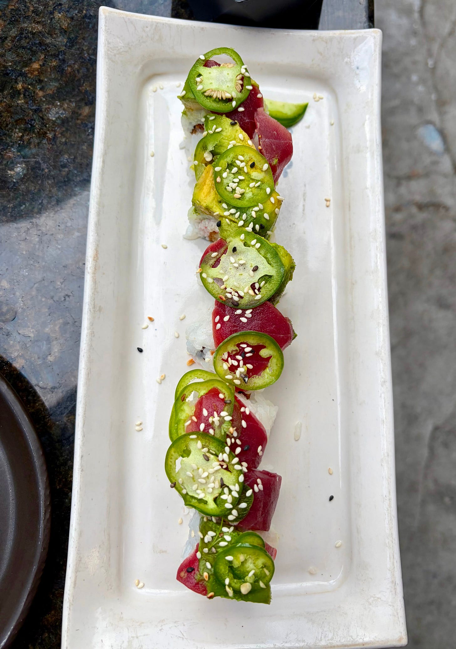 A top-down shot of a gourmet sushi roll arranged vertically on a long, rectangular white ceramic plate. The roll is meticulously topped with alternating layers of deep burgundy raw tuna and bright green avocado slices, crowned with thin, round slices of fresh jalapeño and sprinkled with white sesame seeds.