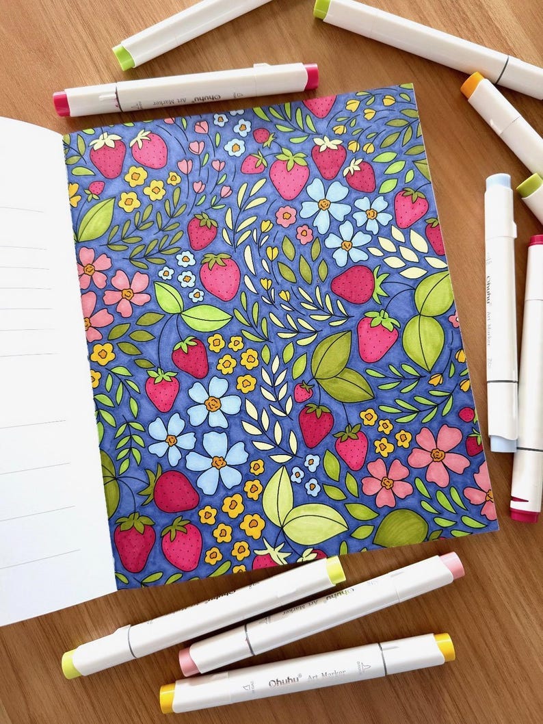 May include: A coloring book page with a pattern of strawberries, flowers, and leaves on a blue background. The page is open to reveal the illustration and is surrounded by several white markers with yellow caps.