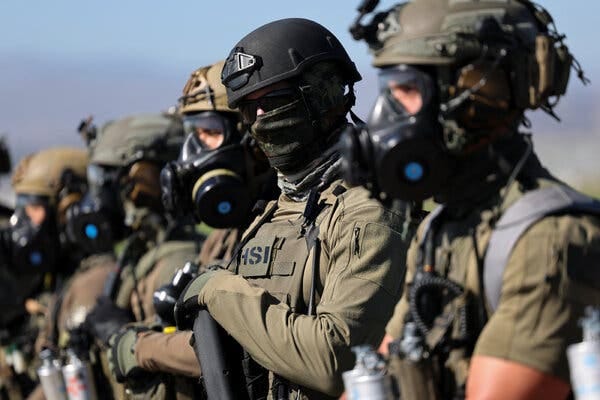 Several agents wearing green uniforms and heavy-duty face masks are seen up close. One of them has a patch that says “HSI,” a reference to Homeland Security Investigations.