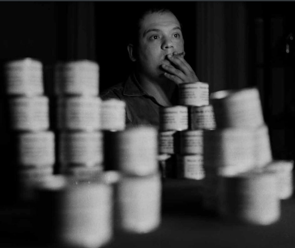 b/w photo of Piero Manzoni smoking, in focus in the background, with some of his tins of shit, roughly piled and out of focus in the foreground