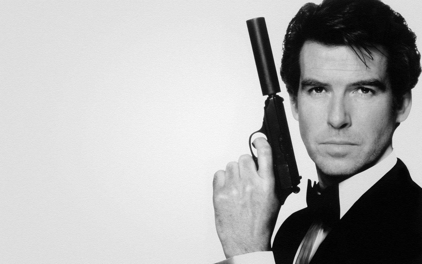 Black and white image of Pierce Brosnan as James Bond, wearing a tuxedo and holding a silenced pistol next to his face, evoking a secret agent aesthetic. Black and white image of Pierce Brosnan as James Bond, wearing a tuxedo and holding a silenced pistol next to his face, evoking a secret agent aesthetic.