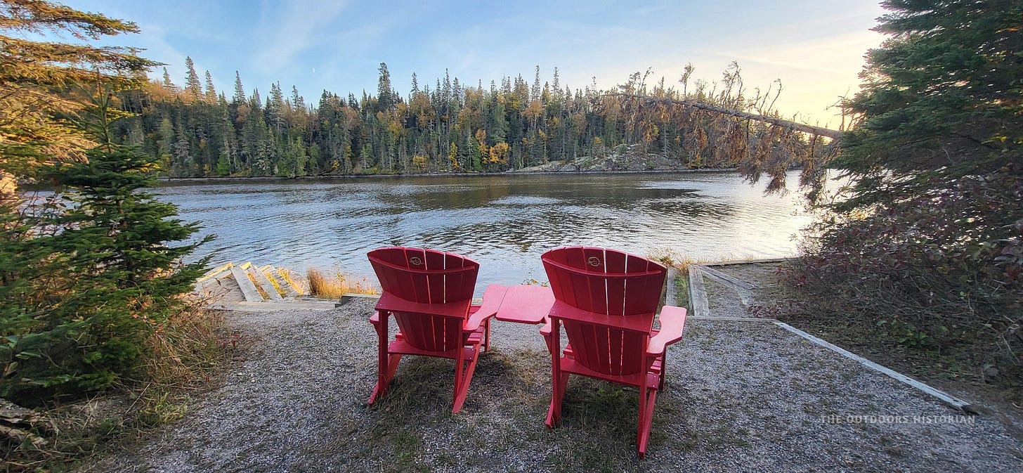 Two bright red Adirondack-style Parks Canada chairs sitting empty on a rocky outcrop. They face a calm, sheltered cove surrounded by a rugged, pine-studded shoreline that looks like a classic Canadian landscape painting.