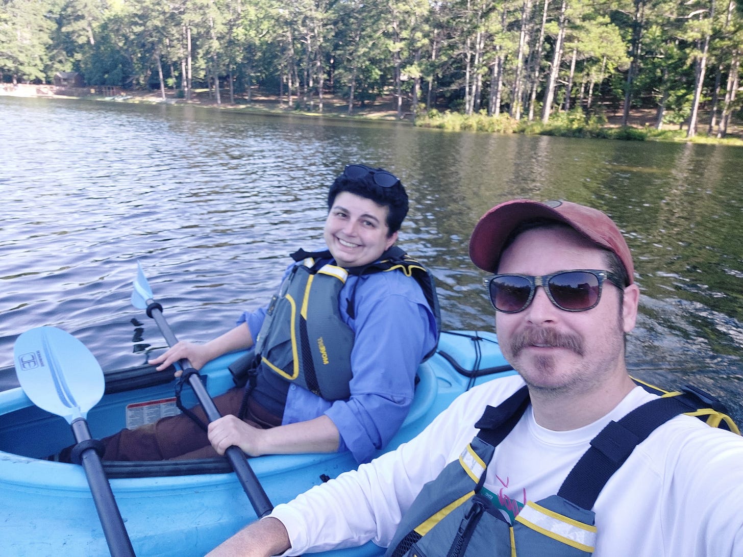 A selfie of two people in separate blue kayaks, side-by-side, floating on a lake on a sunny day. The author is wearing a blue shirt under a blue life jacket and is a white woman in her forties with short dark hair and sunglasses on her head. The photographer, the author's husband, is a white man wearing sunglasses and a white shirt under a blue life jacket. He has medium brown hair under a faded crimson baseball cap and a mustache of the same color.
