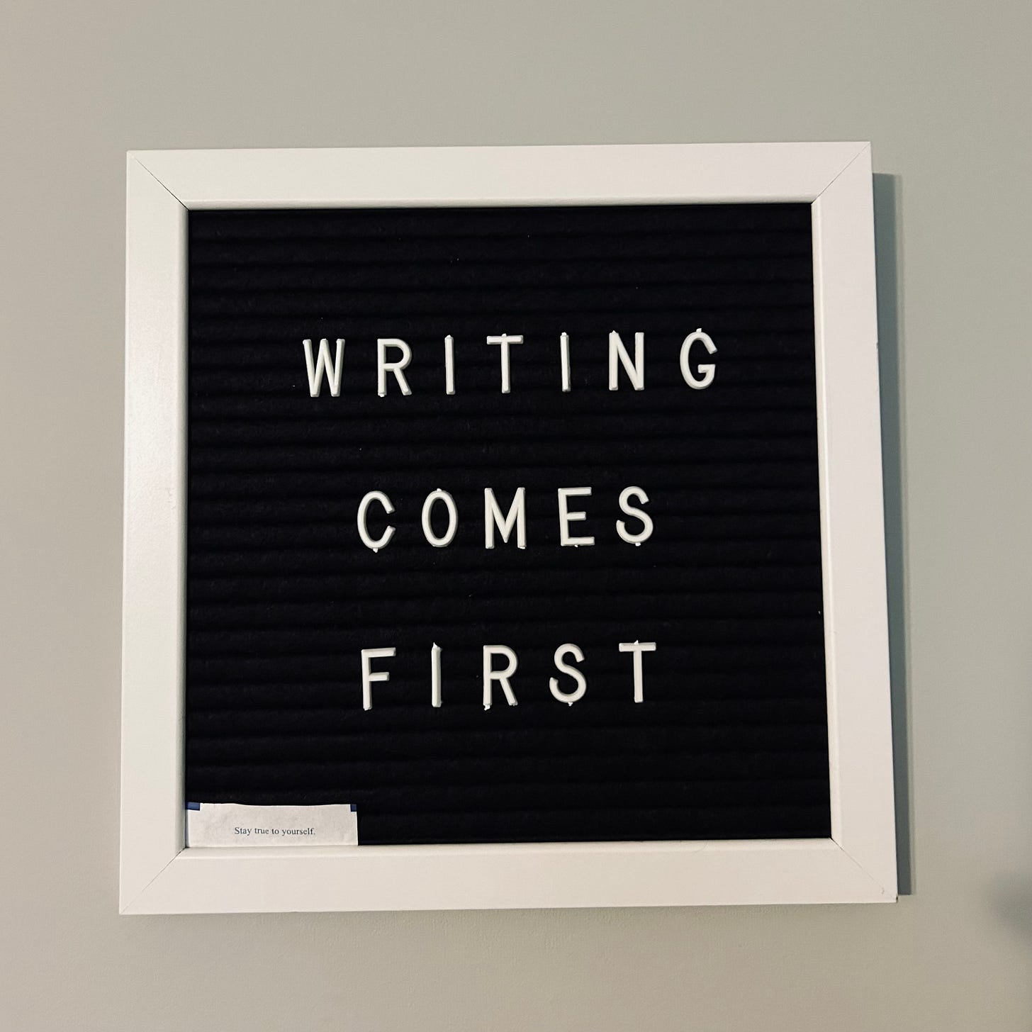 White-framed black felt letter board with the words Writing Comes First spelled out. In the left hand corner there is a small fortune from a fortune cookie that says Stay true to yourself.