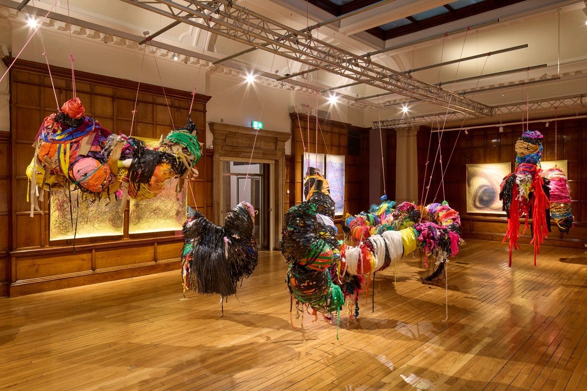 In an art gallery with wood floors and walls, colorful sculptures bound with rope and tape are suspended along with swirling vortex drawings on the walls.