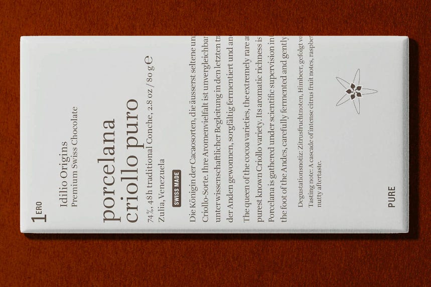 Weisse Schokoladentafel-Verpackung der Schweizer Marke Idilio Origins mit der Aufschrift «porcelana criollo puro – 74 %, 48 h Traditional Conche, Zulia, Venezuela». Die minimalistische Gestaltung zeigt eine Kakaoblüte und den Hinweis «Swiss Made»; die Verpackung liegt auf einem rotbraun-kakaofarbenen Untergrund. (Foto: Jürg Vollmer) Weisse Schokoladentafel-Verpackung der Schweizer Marke Idilio Origins mit der Aufschrift «porcelana criollo puro – 74 %, 48 h Traditional Conche, Zulia, Venezuela». Die minimalistische Gestaltung zeigt eine Kakaoblüte und den Hinweis «Swiss Made»; die Verpackung liegt auf einem rotbraun-kakaofarbenen Untergrund. (Foto: Jürg Vollmer)