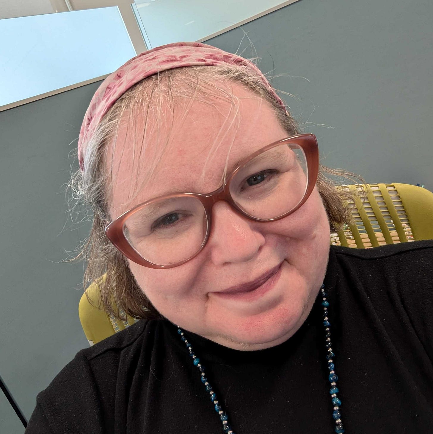 A photo of a woman with "dirty blonde" hair, some gray in the front. She's wearing a black dress and a pink kerchief, and is seated in a brightly-colored chair at the office.