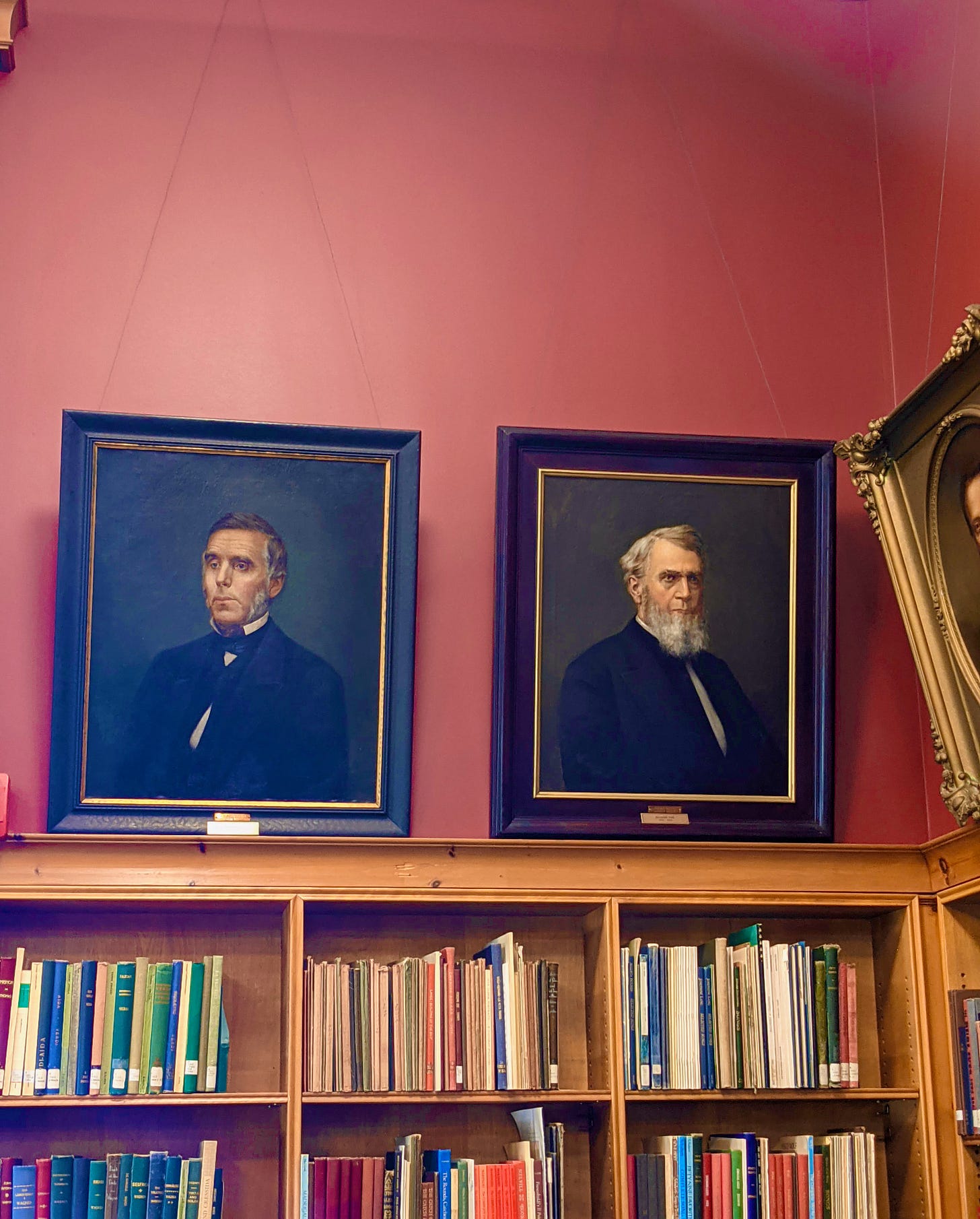A library wall, painted dark pink with several oil portraits of older, white men from the 1800s, in ornate frames. The paintings hang above several shelves of library books.