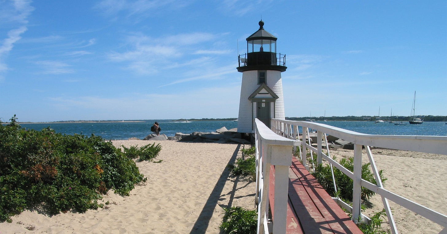 A classic lighthouse with a wooden walkway leading up to it, set against a blue sky and sandy beach with gentle ocean waves in the background.
