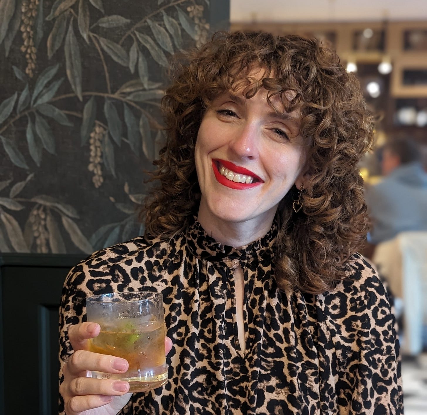 A woman with curly hair and red lipstick smiles and holds up a drink.
