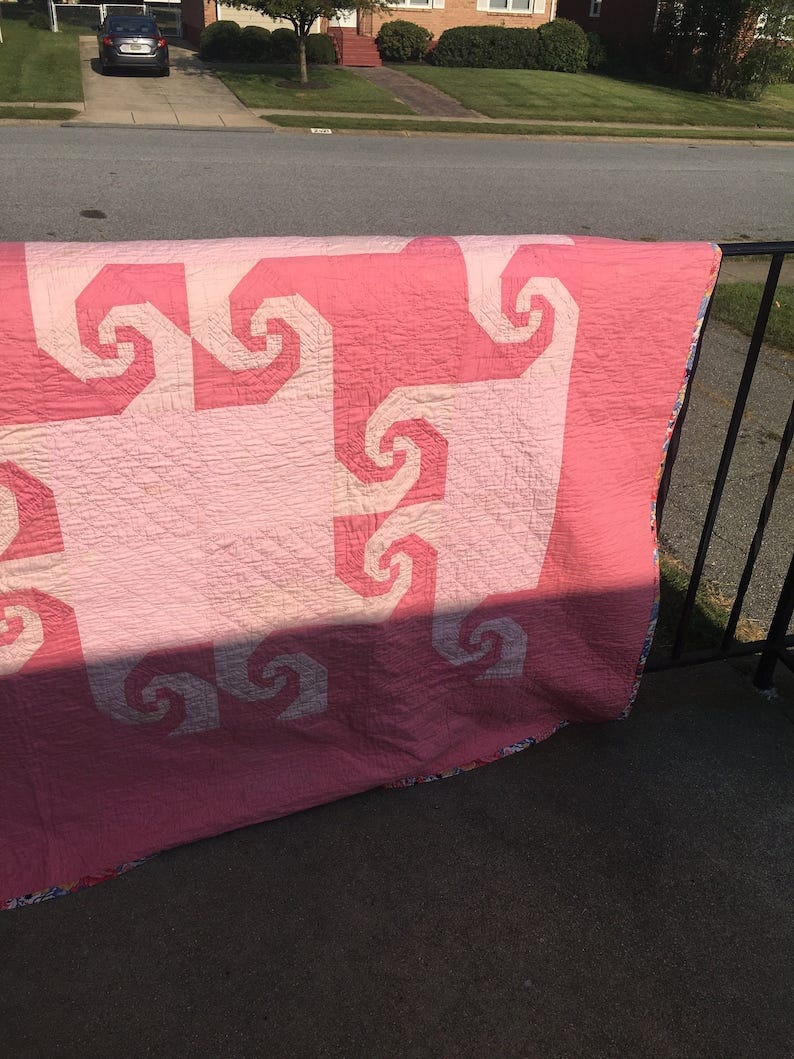 May include: A pink and white quilt with a swirling pattern. The quilt is folded and hanging over a black railing. May include: A pink and white quilt with a swirling pattern. The quilt is folded and hanging over a black railing.