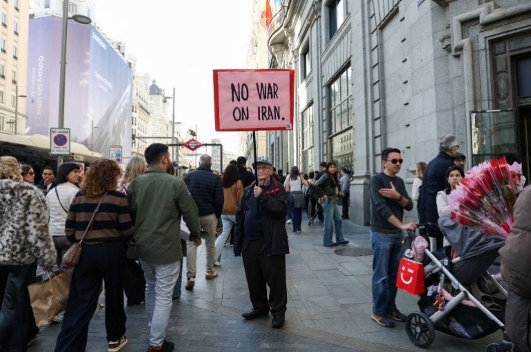 A man on a busy street holds a sign reading 'No war on Iran'. A man on a busy street holds a sign reading 'No war on Iran'.