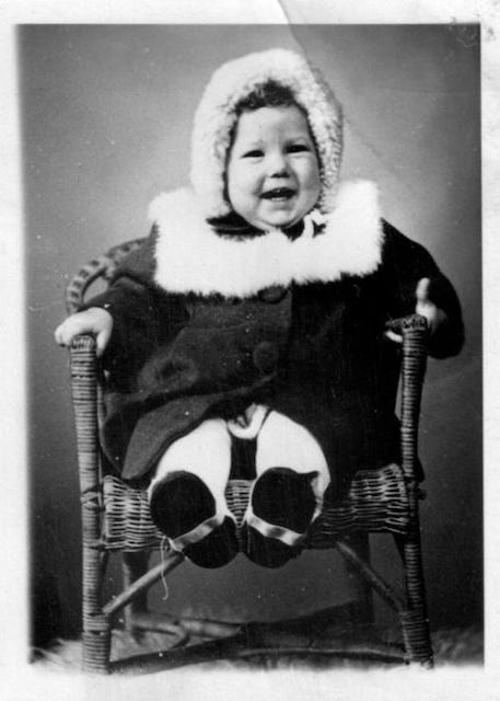 A toddler girl sits in a little wicker chair, feet sticking out from under her, in a dark coat trimmed with white fur, the hood up around her very happy, smiling face, looking at the camera with delight. A toddler girl sits in a little wicker chair, feet sticking out from under her, in a dark coat trimmed with white fur, the hood up around her very happy, smiling face, looking at the camera with delight.