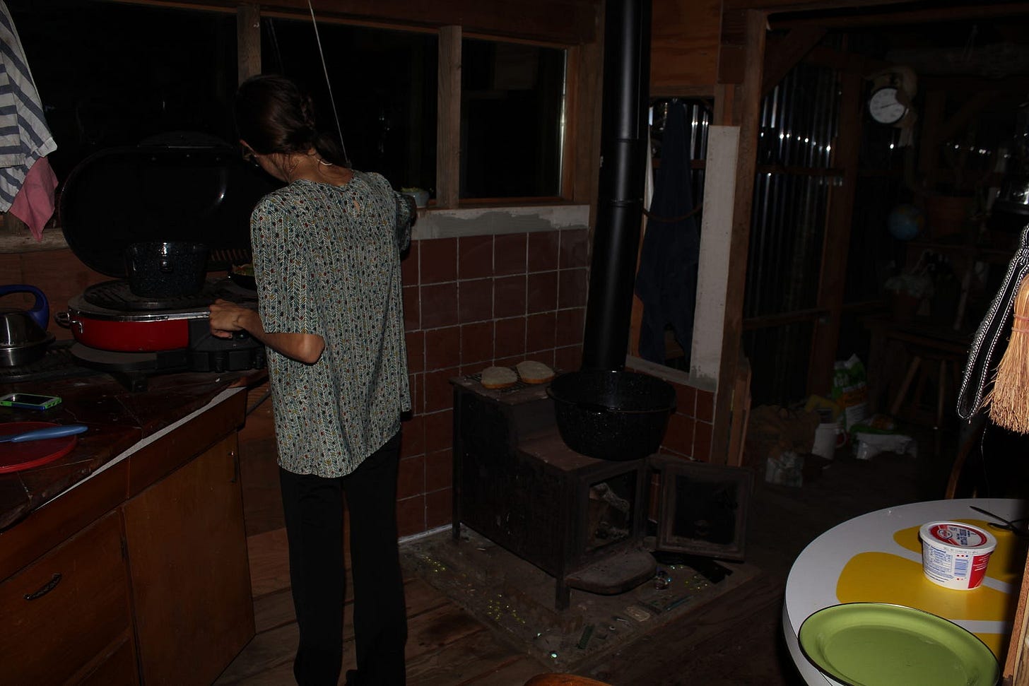 a woman cooking on a camp stove in a dark kitchen with a wood stove next to her