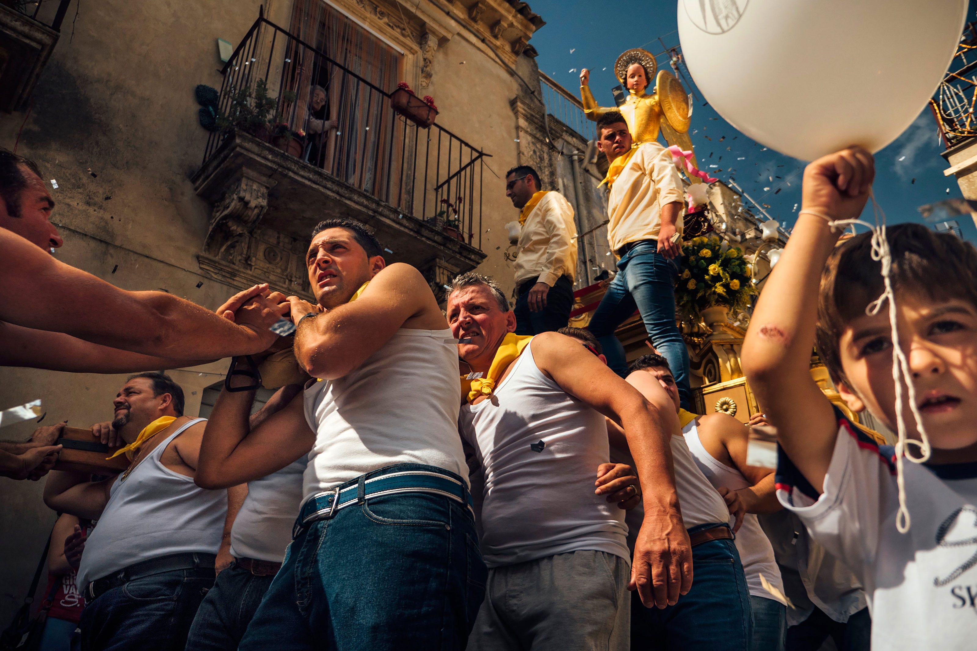 A group of men in white tank tops and yellow scarves carry a religious statue through a crowded street during a lively festival, while a child in the foreground holds a white balloon. Confetti fills the sunny sky.