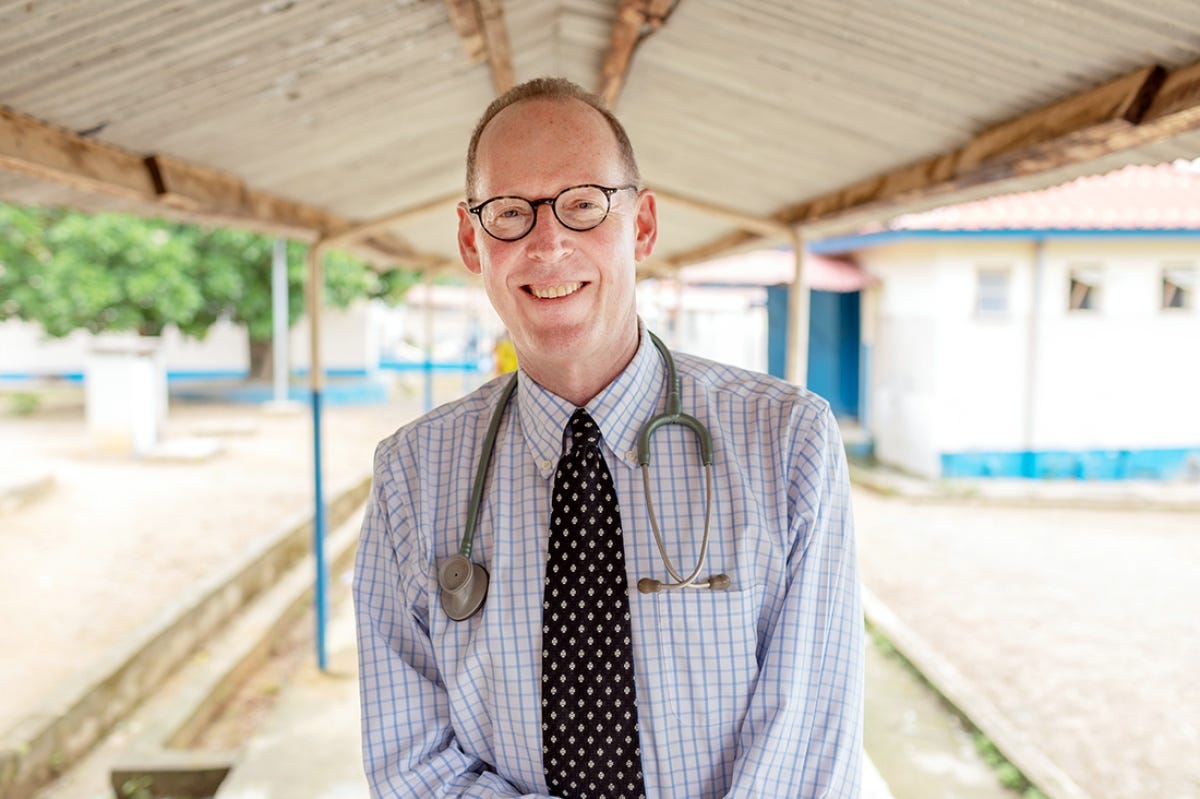 Smiling male doctor with short hair and dark glasses standing in front of a medical clinic.