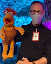 Puppeteer stands looking. atthe camera with an orange fraggle puppet on their arm. Puppeteer stands looking. atthe camera with an orange fraggle puppet on their arm.