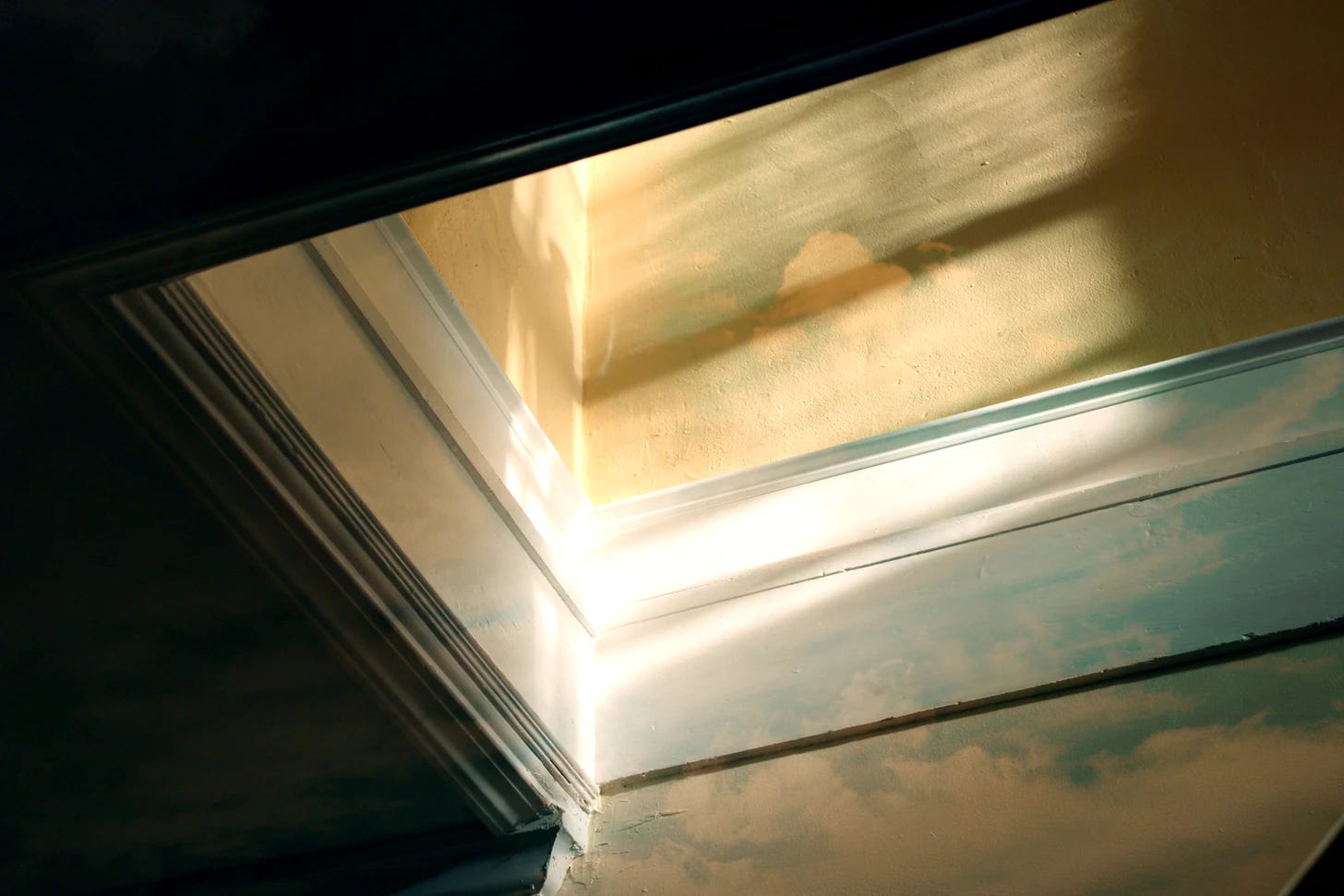 A photo of morning light coming down a staircase. The wall is a light yellow with white moulding and overlays an image of a blue sky with clouds.