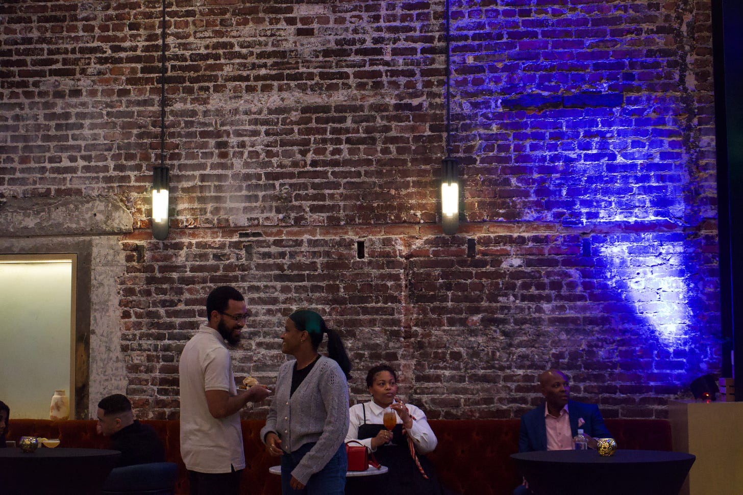 At the same brick-walled venue, a couple stands close together near a red velvet banquette, smiling into each other’s eyes. The woman wears a gray cardigan and dark jeans, her hair pulled back in a ponytail, while the man in a light polo shirt holds a small plate. Behind them, a woman sits at a table with a drink and a red handbag, and a man in a blue suit sits farther down the bench. Warm lighting and blue uplighting illuminate the textured brick wall.