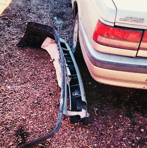 My champagne Mazda 626 lurches cockeyed in the wreck yard with other munched vehicles. The smashed front bumper lies on the ground. The windshield is lanced with cracks. The passenger side is caved in. The drivers side is demolished.
