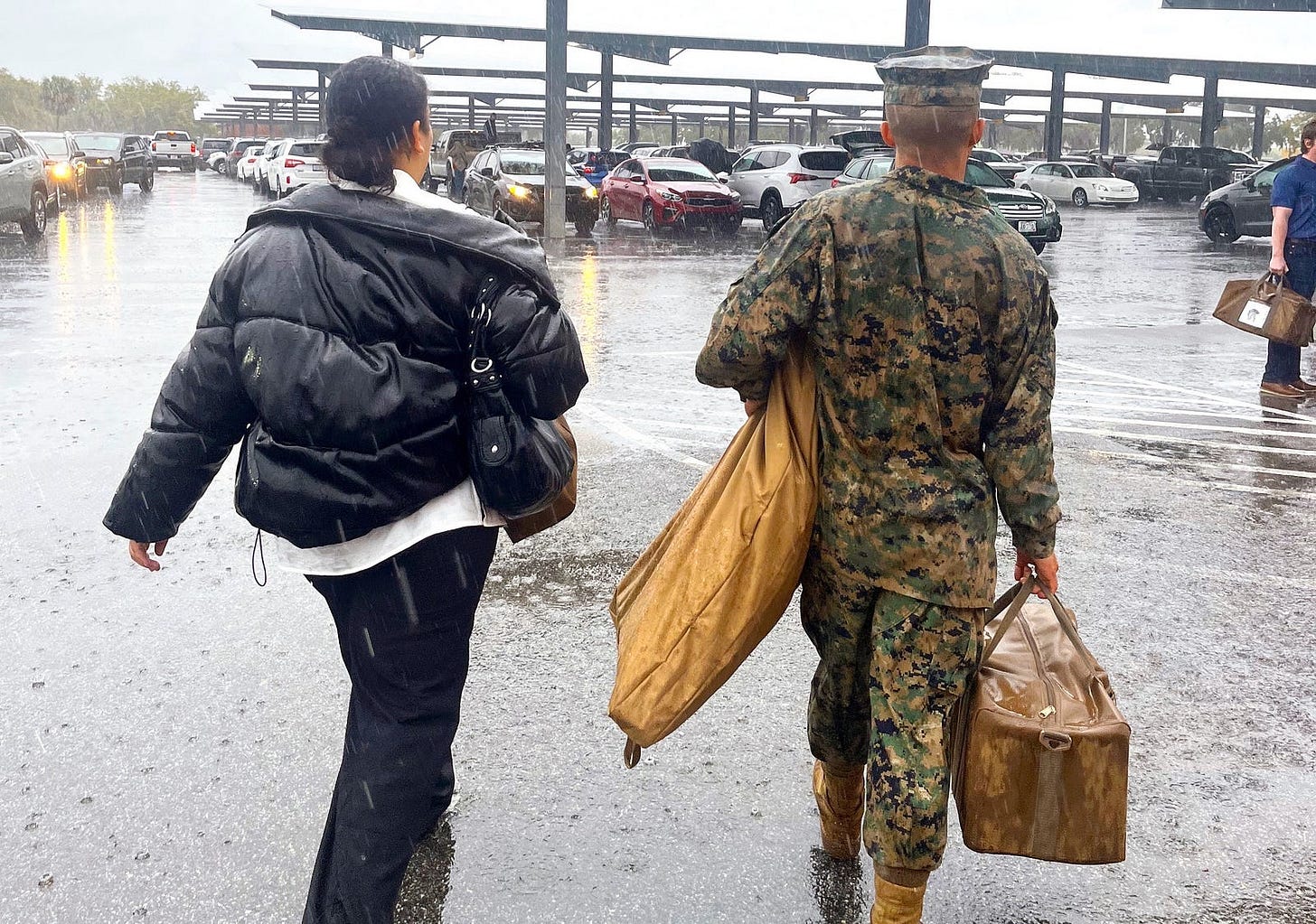 Viewed from behind, through the rain — a young woman in a black jacket walks beside a Marine in camo, carrying bags with his uniform and supplies