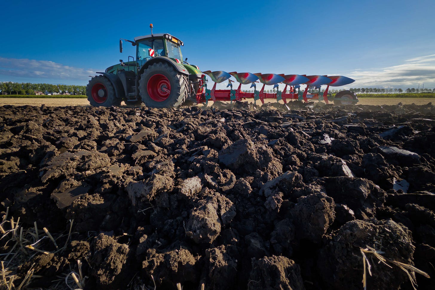 Ein Traktor mit angehängtem Mehrscharpflug pflügt einen Acker; im Vordergrund grobe, frisch gewendete Erdschollen des Oberbodens, im Hintergrund offenes Feld unter blauem Himmel.