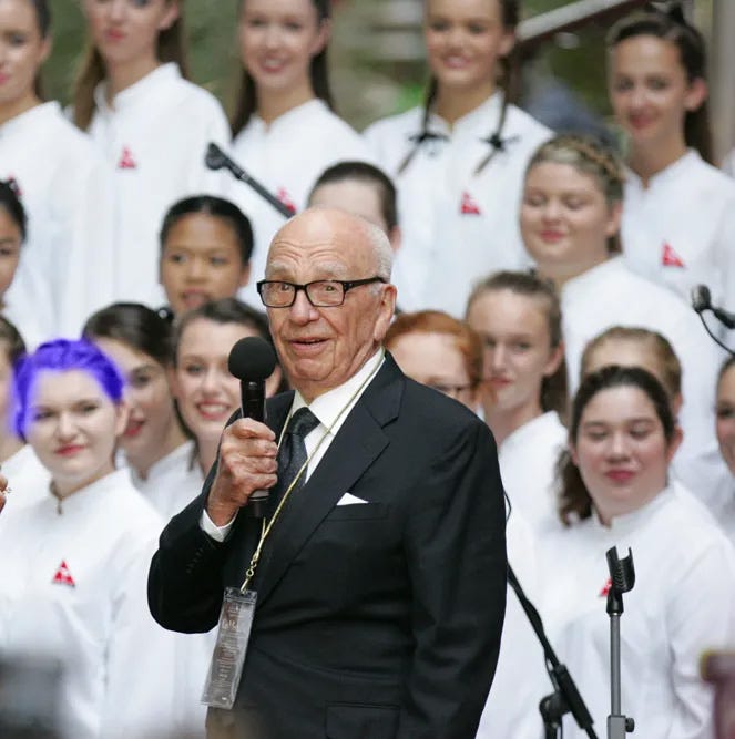 Photo of Rupert Murdoch speaking. A crowd of smiling women is standing behind him, all wearing white, one in blue hair Photo of Rupert Murdoch speaking. A crowd of smiling women is standing behind him, all wearing white, one in blue hair