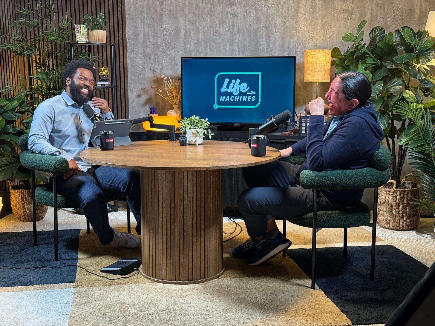 Baratunde Thurston (left) and Michael Running Wolf (right) laughing together while recording an episode of the Life With Machines podcast. They are seated at a round wooden table with microphones, surrounded by plants and a screen displaying the Life With Machines logo in the background Baratunde Thurston (left) and Michael Running Wolf (right) laughing together while recording an episode of the Life With Machines podcast. They are seated at a round wooden table with microphones, surrounded by plants and a screen displaying the Life With Machines logo in the background