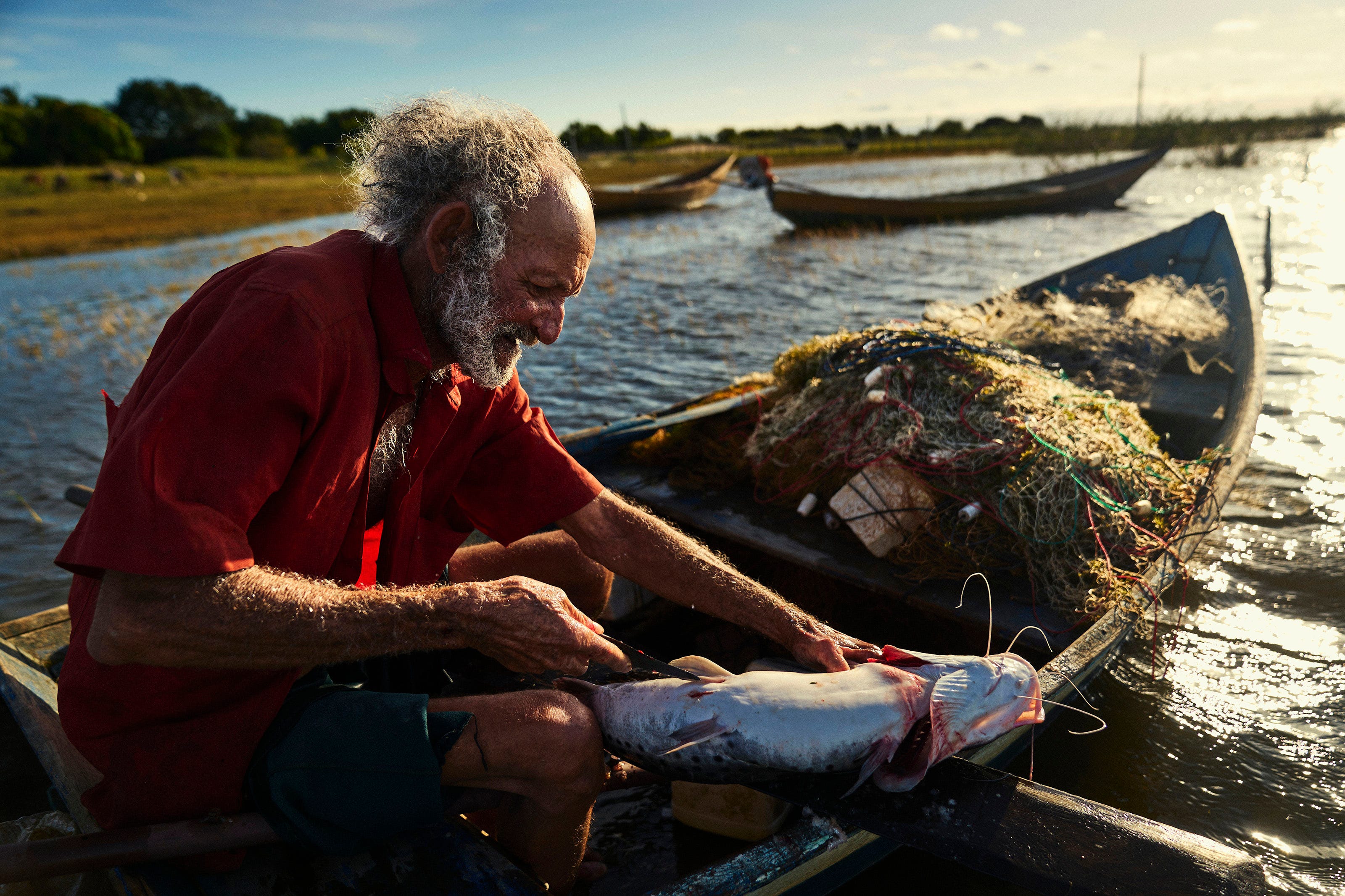 An elderly man with gray hair and a beard sits in a small boat, cleaning a large fish with a knife. Fishing nets and gear are beside him, and the sun sets over the water in the background.