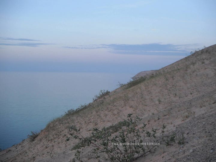 Alt-Text 1: An older photo of the Grand Sable Dunes covered in beach grass. Alt-Text 2: A dusk photo looking down the steep edge of a sand dune toward the water.