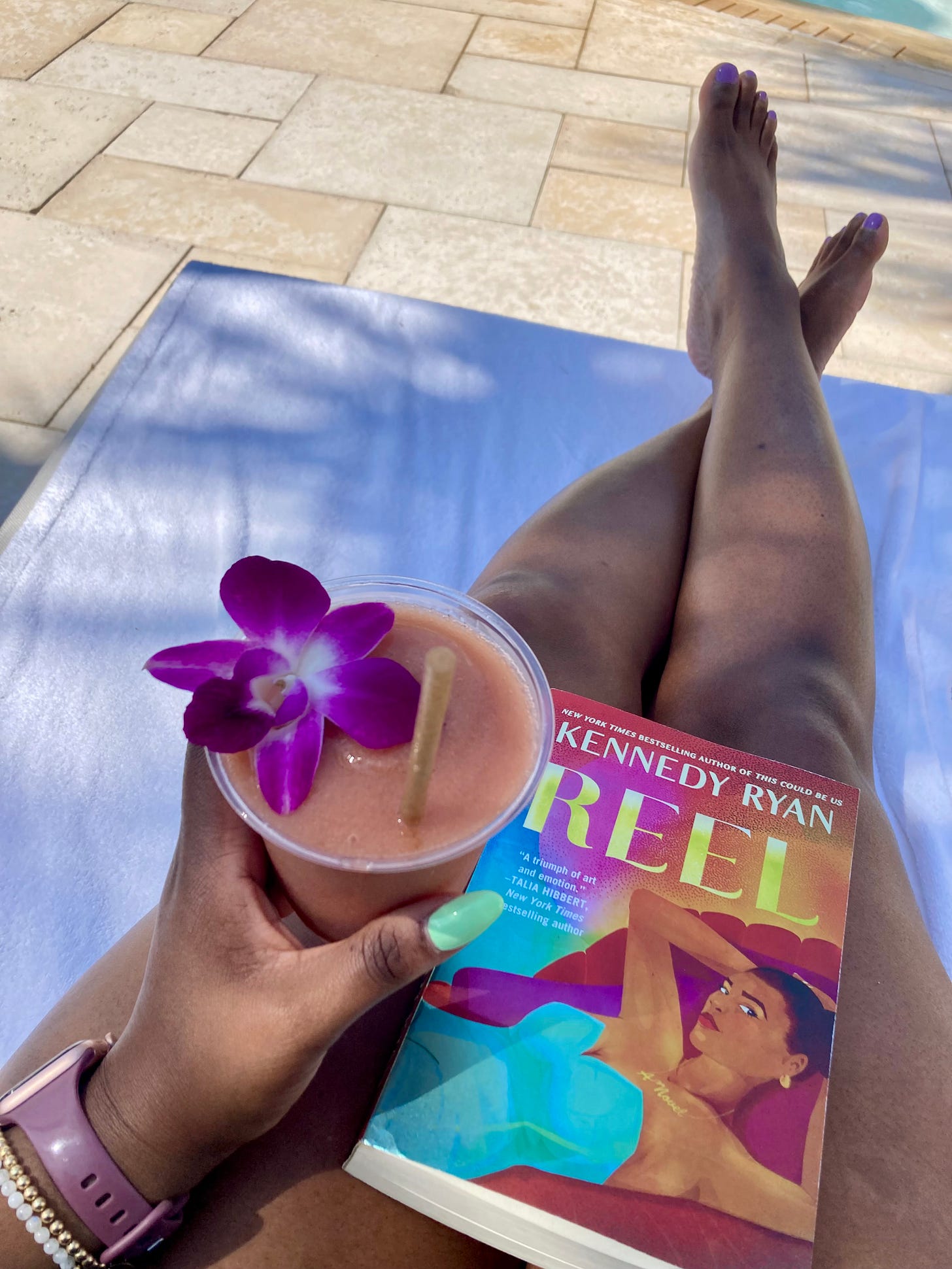 Picture of a Black woman with mint green nails holding a frozen strawberry drink with her legs outstretched and a romance novel in her lap