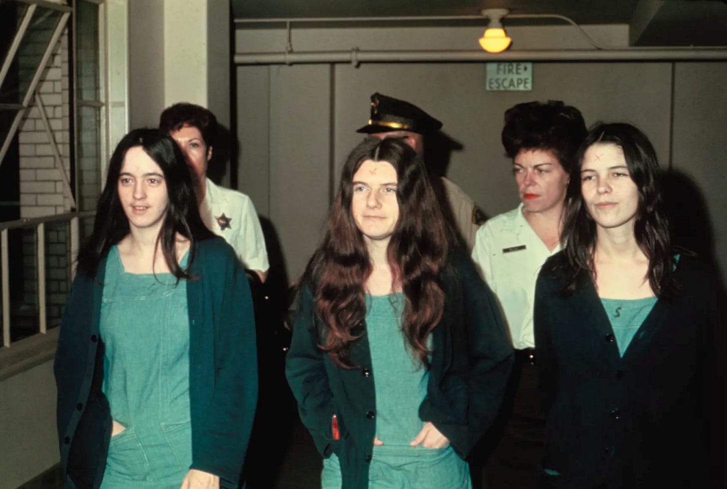 Three white women with red Xs on their foreheads, wearing prison jumpsuits, walk down a corridor followed by three officers Three white women with red Xs on their foreheads, wearing prison jumpsuits, walk down a corridor followed by three officers